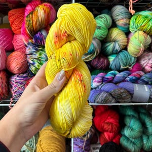 Pu&ograve; includere: Un gomitolo di filato giallo con macchie rosse e arancioni &egrave; tenuto davanti a uno scaffale pieno di vari gomitoli di filato colorati. Il filato &egrave; di un giallo brillante e solare con un motivo maculato, che suggerisce un prodotto tinto a mano o artigianale.
