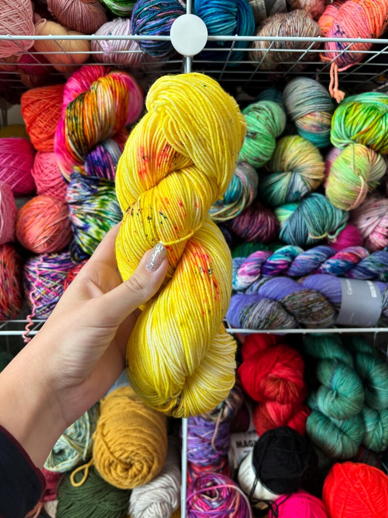 Pu&ograve; includere: Un gomitolo di filato giallo con macchie rosse e arancioni &egrave; tenuto davanti a uno scaffale pieno di vari gomitoli di filato colorati. Il filato &egrave; di un giallo brillante e solare con un motivo maculato, che suggerisce un prodotto tinto a mano o artigianale.