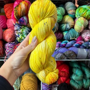 Pu&ograve; includere: Un gomitolo di filato giallo brillante con macchie colorate &egrave; tenuto davanti a uno scaffale pieno di vari gomitoli di filato colorati. Il filato &egrave; di un giallo vibrante con macchie rosse, arancioni e altri colori.