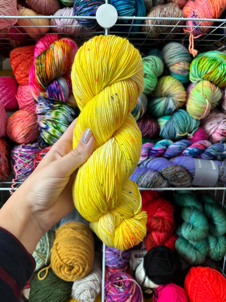 Pu&ograve; includere: Un gomitolo di filato giallo brillante con macchie colorate &egrave; tenuto davanti a uno scaffale pieno di vari gomitoli di filato colorati. Il filato &egrave; di un giallo vibrante con macchie rosse, arancioni e altri colori.