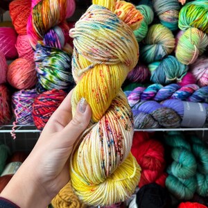 Pu&ograve; includere: Un gomitolo di filato giallo con macchie colorate &egrave; tenuto davanti a uno scaffale pieno di vari colori di filato. Il filato &egrave; giallo brillante con macchie rosse, blu e verdi. Altri colori di filato includono rosa, arancione, verde e viola.
