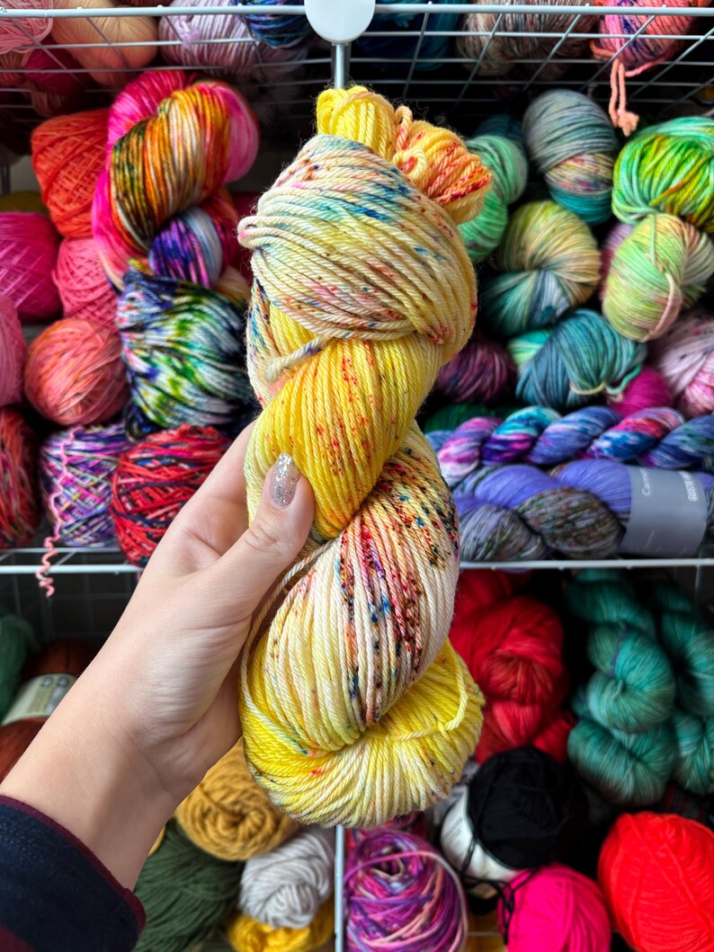 Pu&ograve; includere: Un gomitolo di filato giallo con macchie colorate &egrave; tenuto davanti a uno scaffale pieno di vari colori di filato. Il filato &egrave; giallo brillante con macchie rosse, blu e verdi. Altri colori di filato includono rosa, arancione, verde e viola.