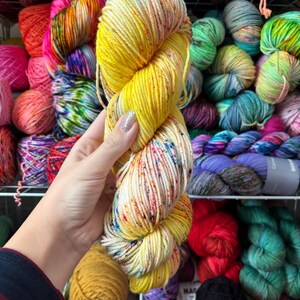 Pu&ograve; includere: Un gomitolo di filato tinto a mano nei toni del giallo e crema, macchiato di blu, arancione e verde. Il filato &egrave; tenuto davanti a uno scaffale pieno di vari gomitoli di filato colorati. Il filato appare morbido e soffice, ideale per progetti di maglia o uncinetto.