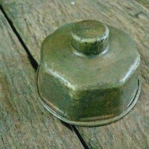 May include: A vintage, tarnished brass container with a rounded top and a small, cylindrical handle. The container has a hexagonal base and sits on a wooden surface. The brass has a greenish hue, suggesting age and use.