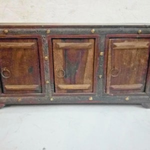May include: A wooden cabinet with three doors, each featuring a ring pull. The cabinet has a dark frame with brass accents and a reddish-brown interior. The doors are a lighter shade of brown, creating a contrast.