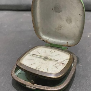 May include: Vintage travel alarm clock with a square face and a green and silver case. The clock face has black numerals and hands. The case is open, revealing the clock's face and the interior of the case. The clock is likely from the mid-20th century.
