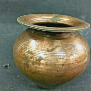 Pode incluir: Um pote antigo de cor cobre com uma borda larga e alargada. O pote tem um corpo arredondado com linhas gravadas horizontais e uma pátina ligeiramente manchada, sugerindo idade e uso. A borda mostra indícios de oxidação verde.