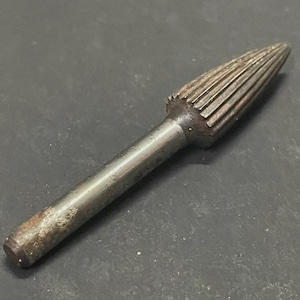 Op de afbeelding: Een metalen roterende frees met een conische kop en een cilindrische schacht. De frees heeft een reeks parallelle groeven op de kop. De tool is gemaakt van metaal en heeft een versleten uiterlijk.