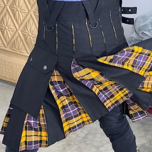 Op de afbeelding: Een zwarte utility kilt met geplooide secties van geel en paars geruite stof. Het kledingstuk is voorzien van een grote cargozak met drukknopen, geïntegreerde D-ringen aan de tailleband en verstelbare zijbanden met metalen drukknopen.