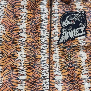 May include: A zip-up jacket with an orange and black tiger stripe pattern. A black patch with a tiger illustration and the word "AVIREX" is on the jacket. The jacket has a brown zipper and collar.