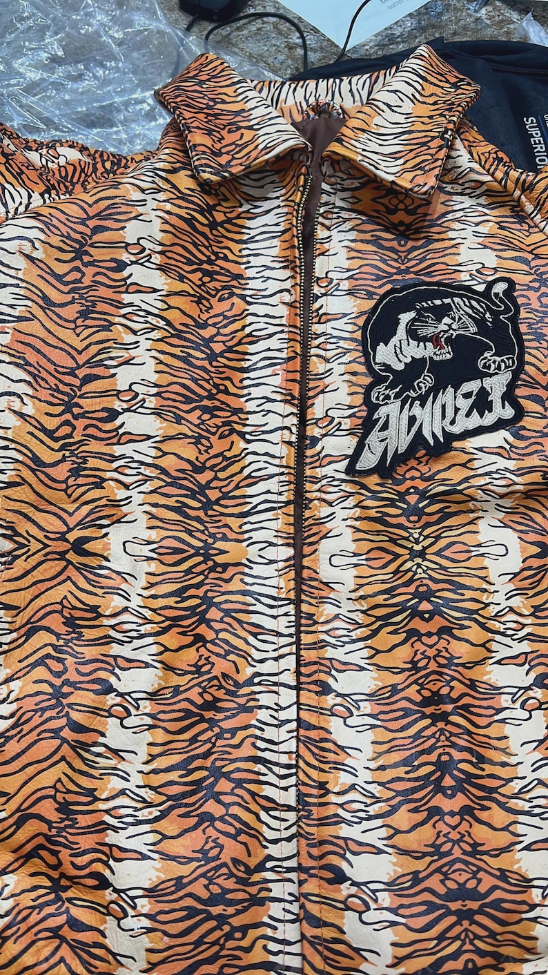 May include: A zip-up jacket with an orange and black tiger stripe pattern. A black patch with a tiger illustration and the word "AVIREX" is on the jacket. The jacket has a brown zipper and collar.