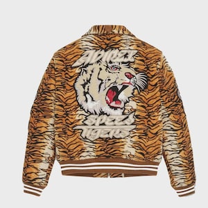 May include: A brown and tan bomber jacket with a tiger stripe pattern. The back of the jacket features a large embroidered tiger head with the text "ADIDAS SPEED TIGERS". The jacket has a brown ribbed collar, cuffs, and hem with white stripes.