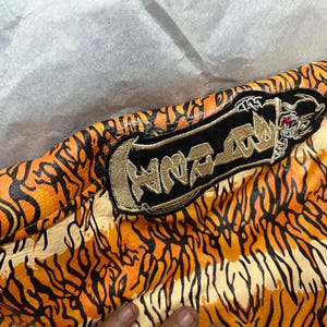 May include: Close-up of a jacket with an orange and black tiger stripe pattern. A black patch with gold embroidery and the word "HNR-RAW" is visible. The jacket's design resembles tiger fur.