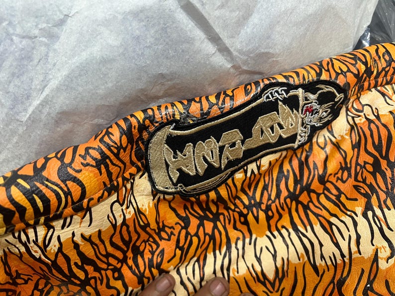 May include: Close-up of a jacket with an orange and black tiger stripe pattern. A black patch with gold embroidery and the word "HNR-RAW" is visible. The jacket's design resembles tiger fur.