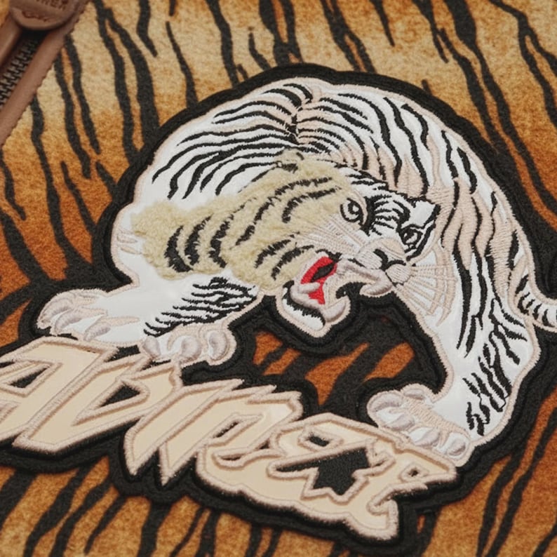 May include: Embroidered patch featuring a snarling tiger with black and white stripes, a red mouth, and beige fur accents. The patch is on a brown and black tiger-striped background. The word "TIGER" is embroidered in beige.