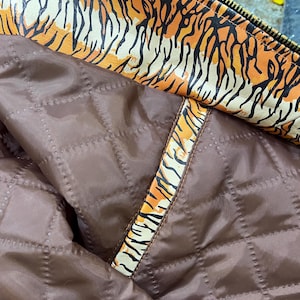 May include: Close-up of a jacket's interior. The lining is a quilted, dark brown material. The trim and collar feature a bold orange and black tiger stripe pattern. A gold zip is visible.
