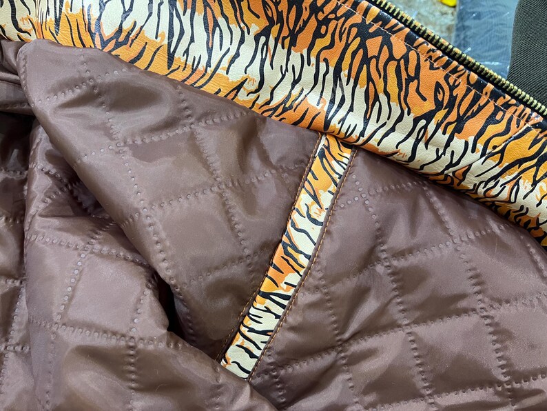 May include: Close-up of a jacket's interior. The lining is a quilted, dark brown material. The trim and collar feature a bold orange and black tiger stripe pattern. A gold zip is visible.