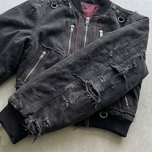 Può includere: Giacca bomber nera invecchiata con multiple cerniere argentate e dettagli ad anello in metallo. La giacca presenta sezioni strappate e strappate, conferendole un'estetica vintage usurata. L'interno è di colore bordeaux scuro.