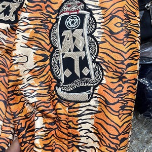 May include: Close-up of a jacket with an orange and black tiger stripe pattern. The jacket has embroidered patches with a tiger and the letters "A.T.". The design of the jacket is reminiscent of a tiger's fur.