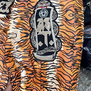 May include: Close-up of a jacket with an orange and black tiger stripe pattern. The jacket has embroidered patches with text and a snake design. The overall aesthetic is bold and eye-catching.