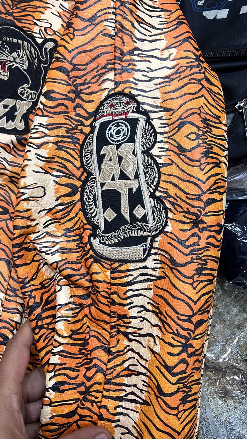May include: Close-up of a jacket with an orange and black tiger stripe pattern. The jacket has embroidered patches with text and a snake design. The overall aesthetic is bold and eye-catching.