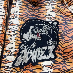 May include: A jacket with a tiger stripe pattern in orange, black, and white. The jacket features a black patch with a panther design and the word "AVIREX" in silver lettering. The jacket has a zipper closure.