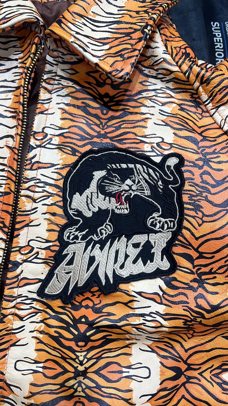 May include: A jacket with a tiger stripe pattern in orange, black, and white. The jacket features a black patch with a panther design and the word "AVIREX" in silver lettering. The jacket has a zipper closure.