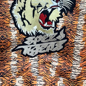 May include: Close-up of a jacket with an orange and black tiger stripe pattern. The jacket has a large embroidered tiger head and the text "Speed Tigers" in silver and black.