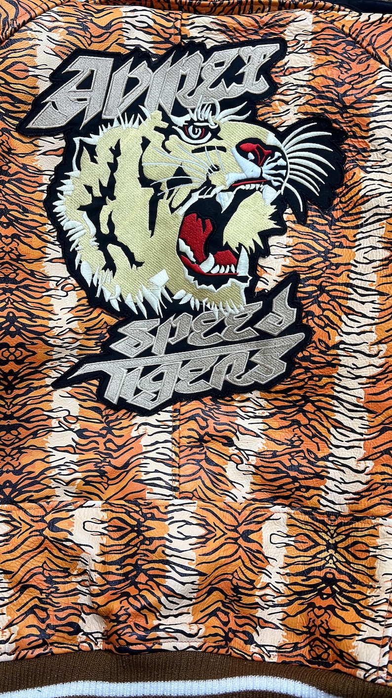 May include: Close-up of a jacket with an orange and black tiger stripe pattern. The jacket has a large embroidered tiger head and the text "Speed Tigers" in silver and black.