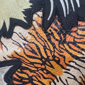 May include: Close-up of a leather patch featuring a tiger stripe design in orange and black. The patch has a cream-coloured background and is bordered by black fabric with white and gold embroidery.