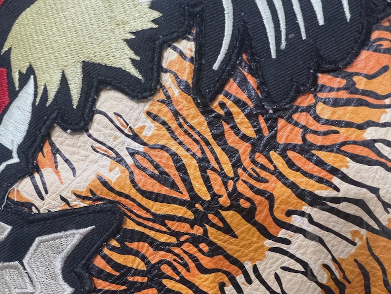 May include: Close-up of a leather patch featuring a tiger stripe design in orange and black. The patch has a cream-coloured background and is bordered by black fabric with white and gold embroidery.