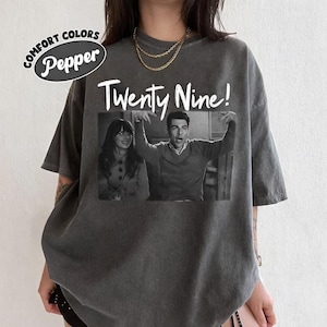 May include: Dark gray t-shirt with the text "Twenty Nine!" above a black and white image of two people. The shirt has a relaxed fit and the words "Comfort Colors Pepper" are printed on the top left.