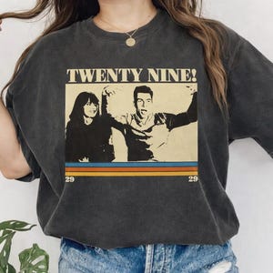 Può includere: T-shirt grigio scuro con grafica retrò. Il testo "TWENTY NINE!" è sopra un'immagine vintage di due persone. Sotto l'immagine ci sono tre strisce orizzontali in blu, arancione e giallo, con il numero "29" su ogni lato.