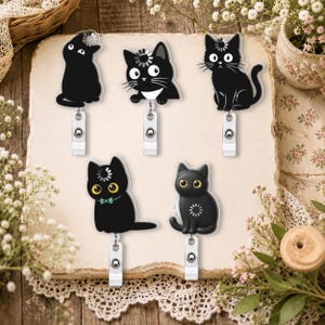 Op de afbeelding: Vijf zwarte kat badgehouders met verschillende houdingen en uitdrukkingen. Elke kat heeft een klein wit laadicoon op het hoofd. De badgehouders zijn gemaakt met een doorzichtige acryl bovenkant en een zilverkleurige metalen clip aan de basis.