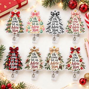 May include: Eight acrylic badge reels shaped like Christmas trees, each featuring the text 'Oh come let us Adore Him' in various fonts. The designs include bows, ornaments, pine branches, and plaid patterns, all attached to clear plastic clips.