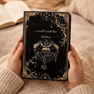 Könnte beinhalten: Schwarzes Notizbuch mit goldenen Akzenten und dem Text "i will not die today". Das Cover zeigt eine Drachenillustration und einen verzierten Rahmen. Im Hintergrund ist ein offenes Buch zu sehen.