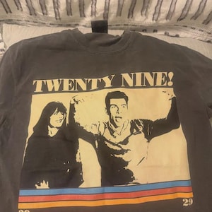 May include: A charcoal gray crew neck t-shirt featuring a graphic print of two people with the text TWENTY NINE! above them. The design includes a beige rectangular background and horizontal stripes in blue, red, and yellow at the bottom with the number 29.