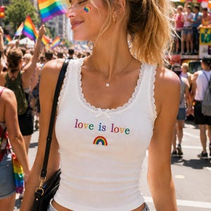 Op de afbeelding: Een wit geribbelde tanktop met een ronde halslijn, afgewerkt met delicaat wit kant en een klein strikje in het midden. Op de borst staat de tekst 'love is love' in veelkleurige letters boven een kleine afbeelding van een regenboog.