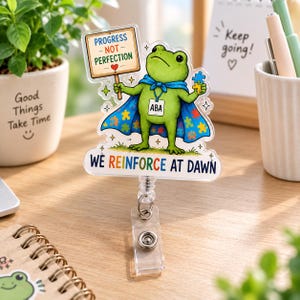 Könnte beinhalten: Ein transparenter Ausweishalter aus Acryl mit einem grünen Frosch, der einen blauen Umhang mit Puzzleteilmuster trägt. Der Frosch hält ein Schild mit der Aufschrift PROGRESS NOT PERFECTION und ein Schild mit ABA. Darunter steht WE REINFORCE AT DAWN.