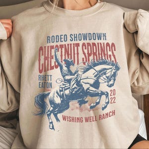 Pode incluir: Camisola bege com um gráfico de rodeio. O design mostra um cowboy montando um cavalo com o texto "Rodeo Showdown Chestnut Springs Rhett Eaton Wishing Well Ranch 2022" em vermelho e azul.