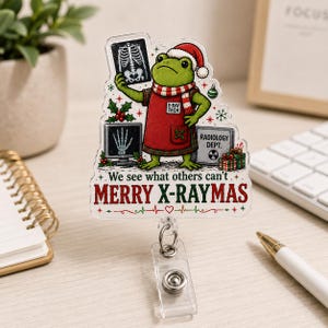 May include: A clear acrylic badge reel featuring a green frog wearing a Santa hat and a red apron labeled X-RAY TECH. The frog holds an X-ray, with text reading We see what others can't and MERRY X-RAYMAS, alongside holiday graphics.
