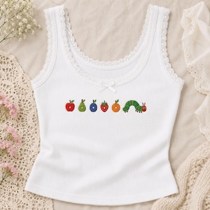 Puede incluir: Una camiseta de tirantes blanca acanalada con escote redondo, ribete de encaje y un pequeño lazo central. El pecho presenta un bordado con una manzana roja, una pera verde, una ciruela azul, una fresa, una naranja y una oruga verde.