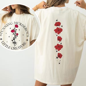 Op de afbeelding: Crèmekleurig T-shirt met een cirkelvormige afbeelding op de voorkant met de tekst "Stunning Murderous Little Creature". De afbeelding toont een roos en een slang. De achterkant van het shirt heeft een verticaal ontwerp van rode bloemen.
