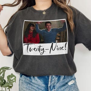 Può includere: T-shirt grigio scuro con un'immagine incorniciata di due persone e la scritta "Twenty-Nine!". L'immagine mostra una donna con un cappotto rosso e un uomo con un maglione blu. La t-shirt è uno stile casual.