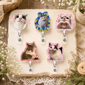Op de afbeelding: Vijf intrekbare badgehouders met verschillende kattenmemes. De bovenste rij toont een kat met een roze strik, een kat op een blauwe sterrenachtergrond met de tekst HUH, en een kat die een hartje vormt. De onderste rij toont een verraste en een beleefde kat.