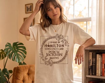 May include: A cream-coloured short-sleeve t-shirt featuring a dark brown graphic print. The design includes a circular border with floral accents and the text: I SPEAK IN HAMILTON SONG LYRICS AND PERCY JACKSON QUOTES, with a star and trident icon.