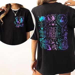 Può includere: T-shirt nera con un disegno a forma di cuore sul davanti e una grafica più grande sul retro. Il disegno frontale presenta un arciere e il testo "Archer". La grafica sul retro include il testo "Once Upon a Broken Heart" e altre illustrazioni in verde acqua e viola.