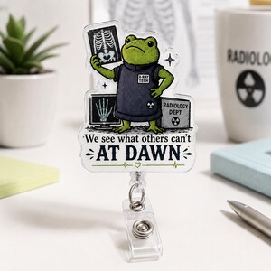 May include: A clear acrylic badge reel featuring a green frog dressed as an X-ray technician, holding a skeletal X-ray film. The design includes a monitor, a box labeled RADIOLOGY DEPT, and the text We see what others can't AT DAWN.