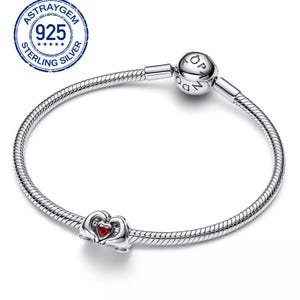 May include: A sterling silver bracelet with a heart-shaped charm. The charm features a red heart-shaped stone. The bracelet has a snake chain design and a round clasp with the word "Pandora" on it. The image also includes a "925 Sterling Silver" stamp.