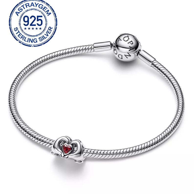 May include: A sterling silver bracelet with a heart-shaped charm. The charm features a red heart-shaped stone. The bracelet has a snake chain design and a round clasp with the word "Pandora" on it. The image also includes a "925 Sterling Silver" stamp.